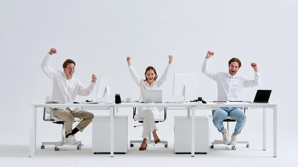 Three colleagues at a bright, uncluttered shared desk, celebrating at the end of a smooth workday